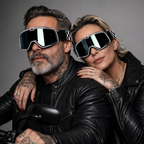 A man and a woman wearing oversized black and silver motocross-style goggles with mirrored lenses, posed together in black leather jackets against a neutral gray studio background.