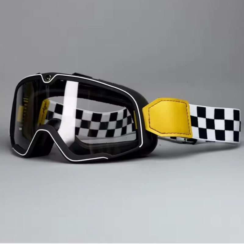 Black Motorcycle goggles with a clear lens, white trim, a yellow strap connector, and a black-and-white checkered strap, shown on a neutral gray background.
