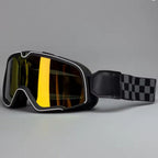 Black Motorcycle goggles with a reflective yellow-tinted lens, white trim, and a black-and-white checkered strap, shown on a neutral gray background.