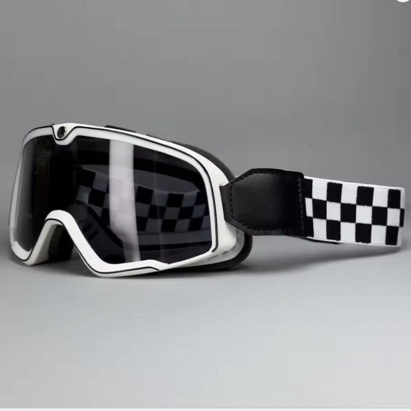 A pair of Motorcycle goggles with a white frame, dark tinted lenses, and a black and white checkered strap, shown on a neutral gray background.