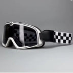A pair of Motorcycle goggles with a white frame, dark tinted lenses, and a black and white checkered strap, shown on a neutral gray background.