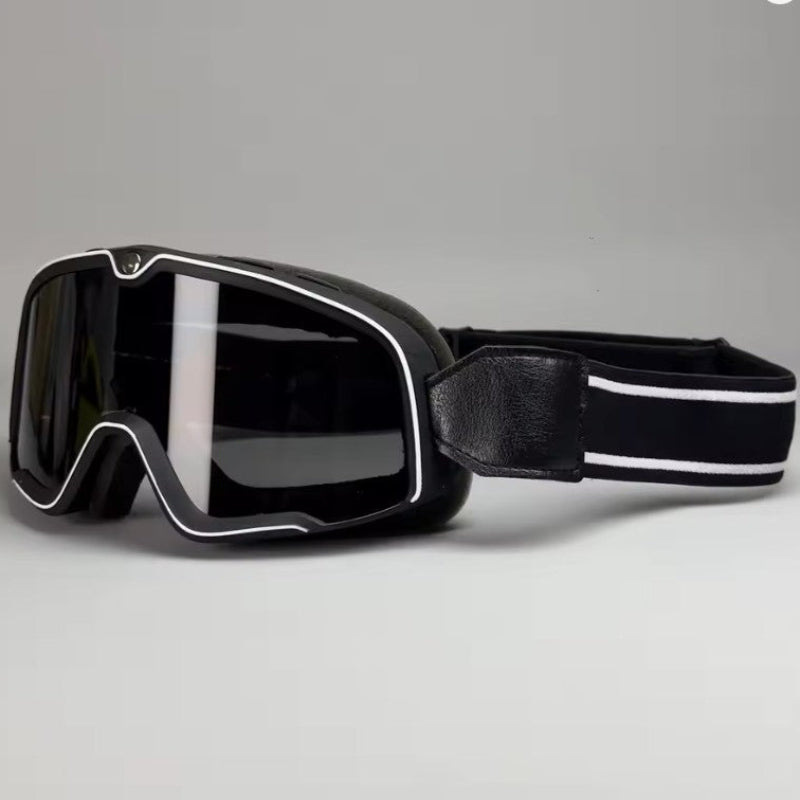 Black Motorcycle goggles with a wide dark lens, silver trim, and a black adjustable strap with white stripes, shown on a neutral gray background.