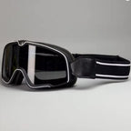Black Motorcycle goggles with a wide dark lens, silver trim, and a black adjustable strap with white stripes, shown on a neutral gray background.