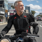 A man wearing a black motorcycle riding jacket with red accents sits on a motorcycle on a city street.
