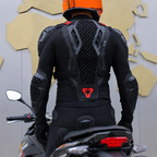 A person wearing a black full-body motorcycle riding suit with integrated protective armor and an orange helmet, photographed from behind while seated on a motorcycle. The suit features a honeycomb mesh back panel and a red geometric logo on the lower back.