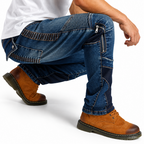 A man wearing blue denim work pants with distinctive ribbed knee patches, contrast stitching, and zippered pockets, paired with brown suede work boots and a white t-shirt, shown in a crouching pose against a white studio background.