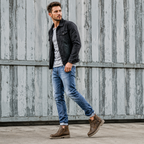 A man wearing a black denim jacket, light gray t-shirt, blue jeans, and brown boots, standing in front of a weathered gray corrugated metal wall.