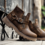 A pair of dark brown leather ankle boots with buckle straps and rugged soles, resting on a weathered wooden surface against a rustic metal gate.
