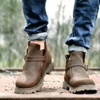 A person wearing brown leather ankle boots with elastic side panels and buckle straps, paired with cuffed blue jeans, standing outdoors on a wooden surface with a blurred natural background.