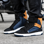 A close-up of a person wearing a pair of high-top sneakers with black, navy blue, and tan panels, paired with black jeans, standing on a gray concrete surface.