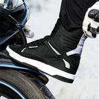 A close-up of a person wearing a black and white high-top motorcycle boot, with a gloved hand holding the ankle, next to a motorcycle tire.