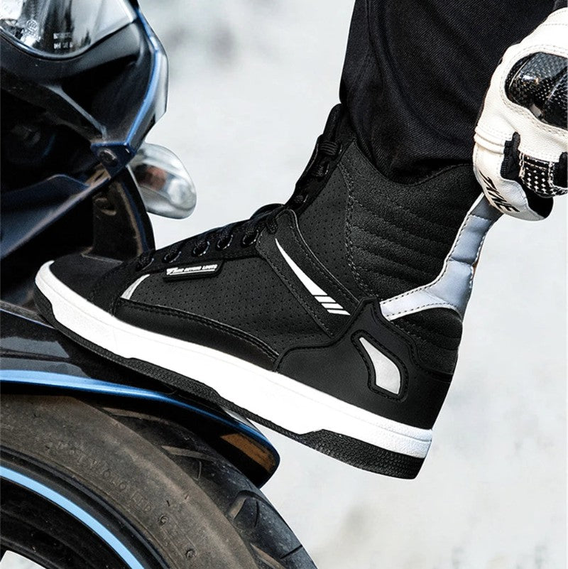 A close-up of a person wearing a black and white high-top motorcycle boot, with a gloved hand holding the ankle, next to a motorcycle tire.