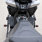 Close-up rear view of a motorcycle showing the seat, rear rack, foot pegs, and front section of the bike in black and silver on a light gray paved surface.