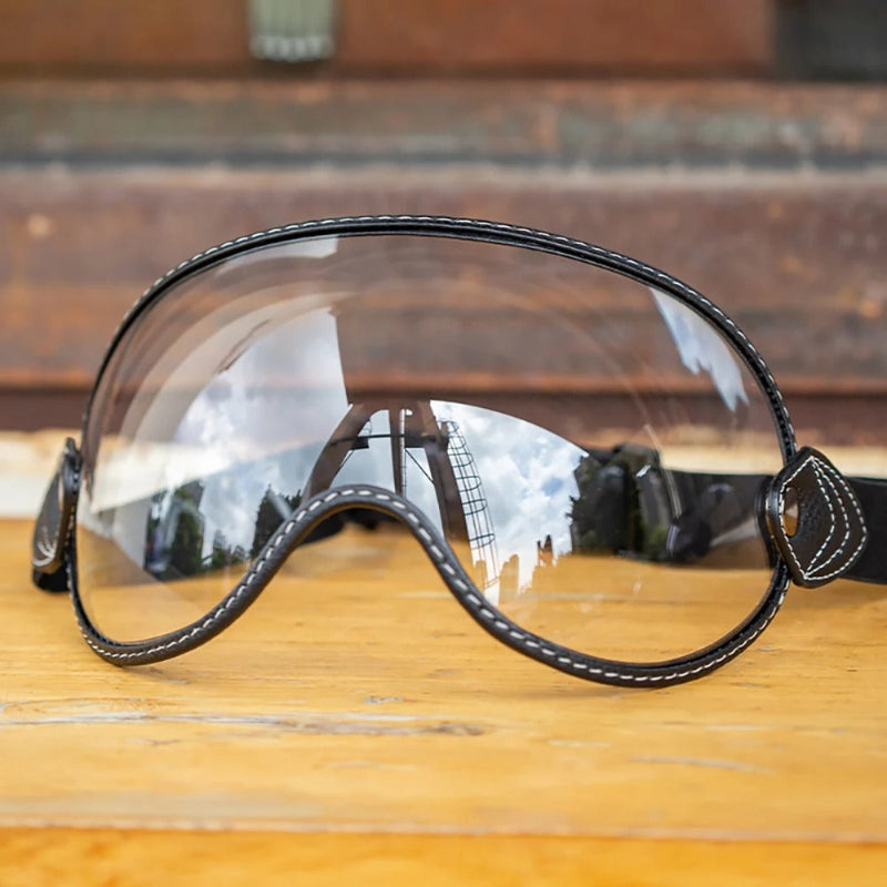 Clear protective goggles with a black stitched frame and strap, resting on a wooden surface with a rustic brown background.