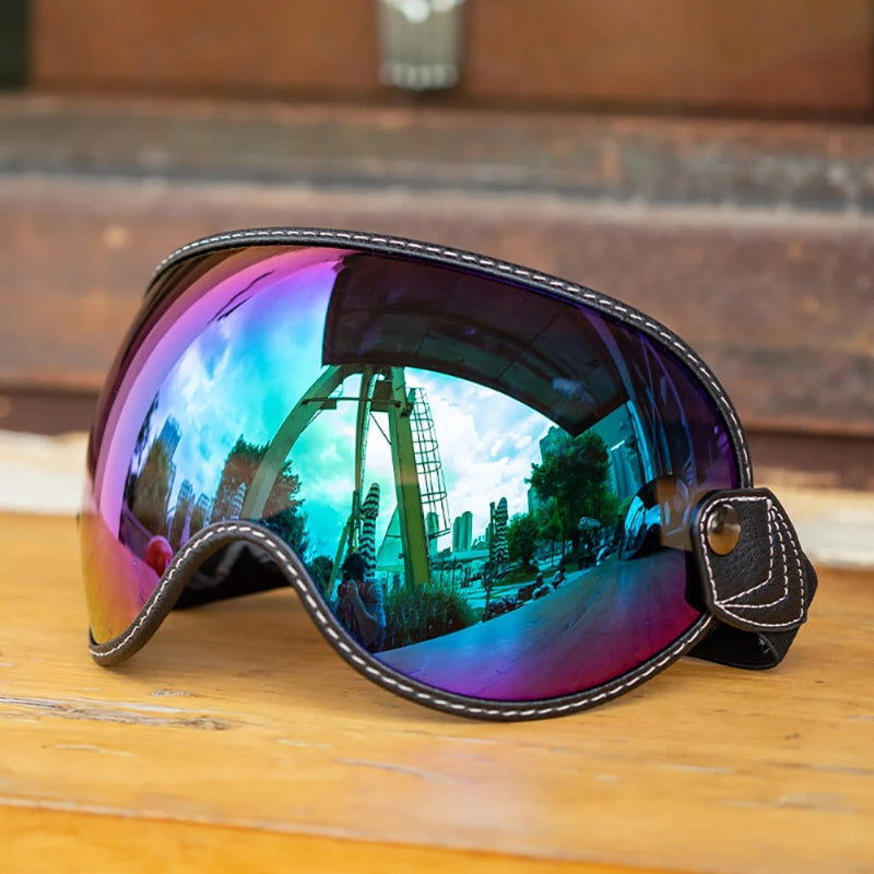 A pair Motorcycle-Goggles with a large curved shield lens, featuring a black leather frame with white contrast stitching and a matching strap. The lens has an iridescent, color-shifting finish reflecting blue, purple, and pink hues, with a warm outdoor scene reflected inside.