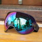 A pair Motorcycle-Goggles with a large curved shield lens, featuring a black leather frame with white contrast stitching and a matching strap. The lens has an iridescent, color-shifting finish reflecting blue, purple, and pink hues, with a warm outdoor scene reflected inside.