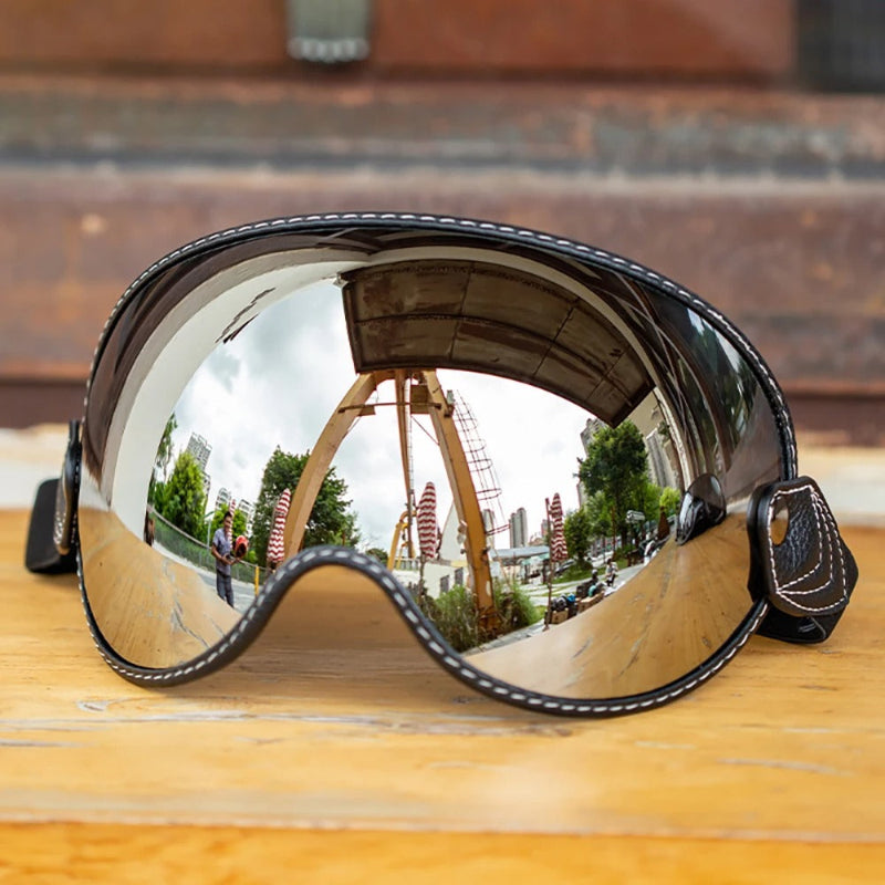 Large shield-style Motorcycle-Goggles with a mirrored reflective lens, black frame, white stitching, and adjustable straps, resting on a wooden surface with an outdoor scene reflected in the lens.