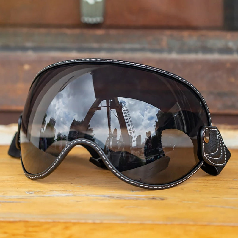 Black Motorcycle-Goggles with white contrast stitching and dark mirrored lenses, resting on a wooden surface with a blurred outdoor background.