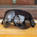 Black Motorcycle-Goggles with white contrast stitching and dark mirrored lenses, resting on a wooden surface with a blurred outdoor background.
