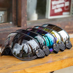 five motorcycle goggles with dark leather straps and colorful mirrored lenses, arranged in a row on a wooden surface.