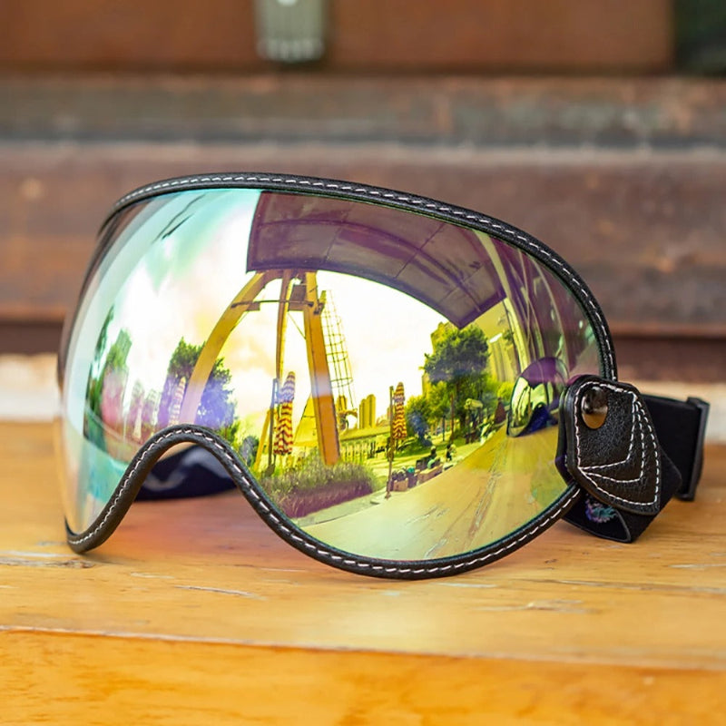 A pair of Motorcycle-Goggles with a large curved reflective lens, black leather trim with white stitching, and an adjustable strap, resting on a wooden surface.