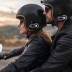 Two people wearing black helmets with mounted communication devices, riding a motorcycle outdoors during sunset.