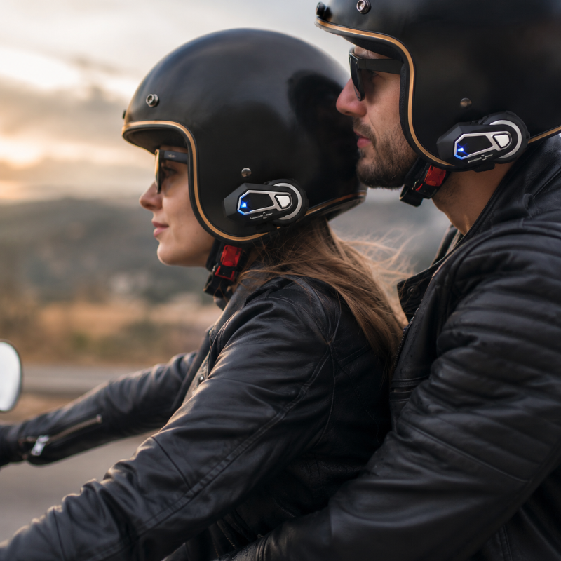 Two people wearing black helmets with mounted communication devices, riding a motorcycle outdoors during sunset.