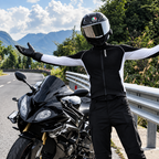 A person wearing a black and white motorcycle riding suit, black gloves, and a black helmet stands with arms outstretched next to a black sport motorcycle on a road with mountains in the background.