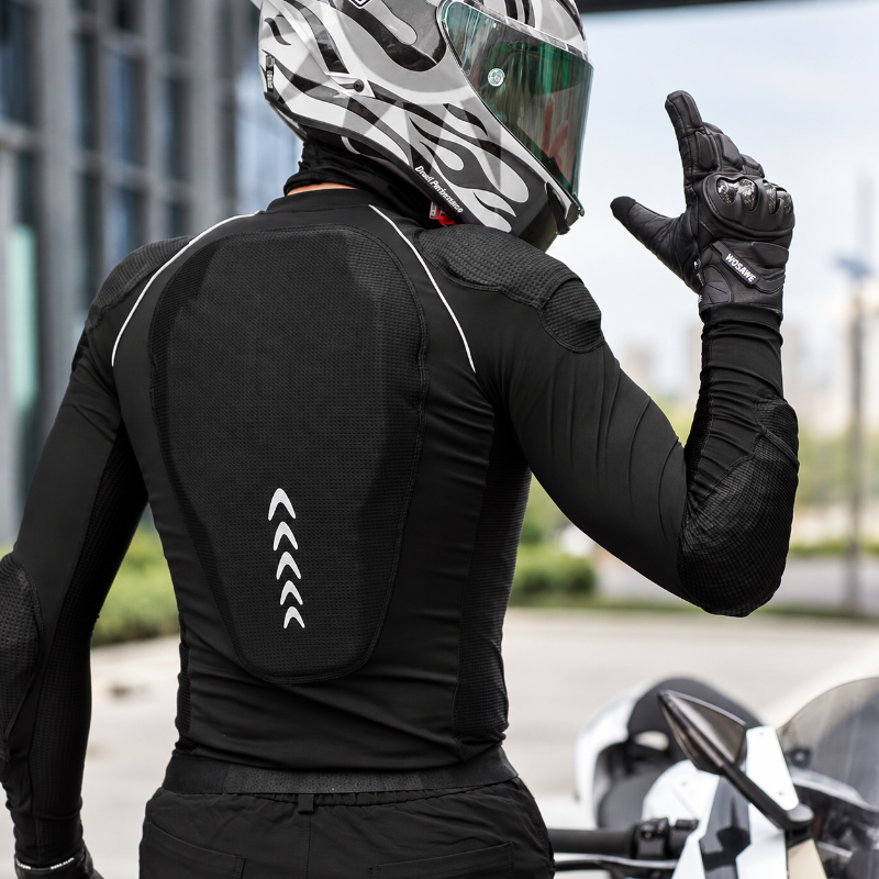 A person wearing a black motorcycle riding jacket with integrated back armor, black gloves, and a white, gray, and black full-face helmet, photographed outdoors with a blurred motorcycle and city background.