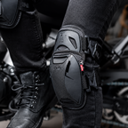 Close-up of a rider's knee wearing a black motorcycle knee protector with adjustable straps and a small red brand tab, paired with black pants and boots, with a blurred motorcycle in the background.