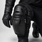 Black motorcycle knee protector with integrated gloves and pants, shown on a model in a crouching position against a light gray studio background.