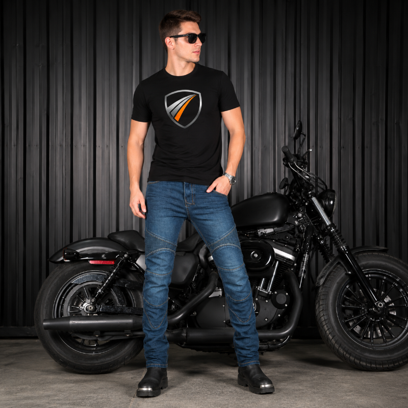 A man wearing a black t-shirt  with a metallic shield Bonny's  logo on the chest, blue jeans, and sunglasses stands in front of a black cruiser motorcycle against a dark corrugated metal wall.