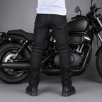A person wearing black denim motorcycle pants with contrast gold stitching stands in front of a black cruiser motorcycle on a gray studio background.