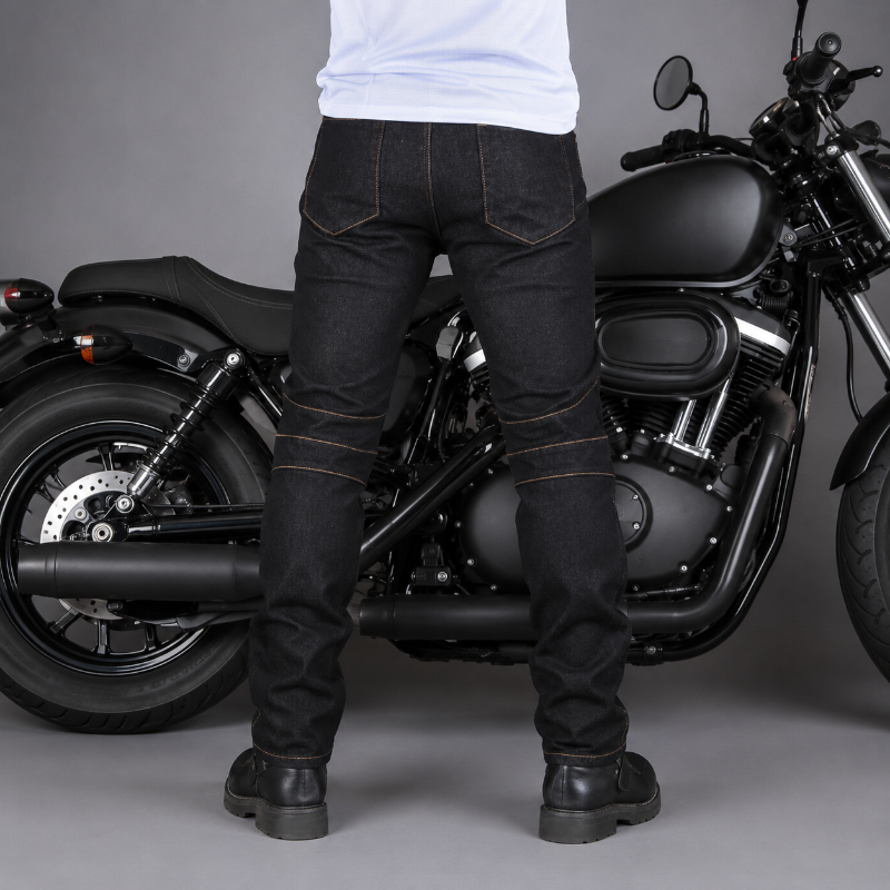 A person wearing black denim motorcycle pants with contrast gold stitching stands in front of a black cruiser motorcycle on a gray studio background.