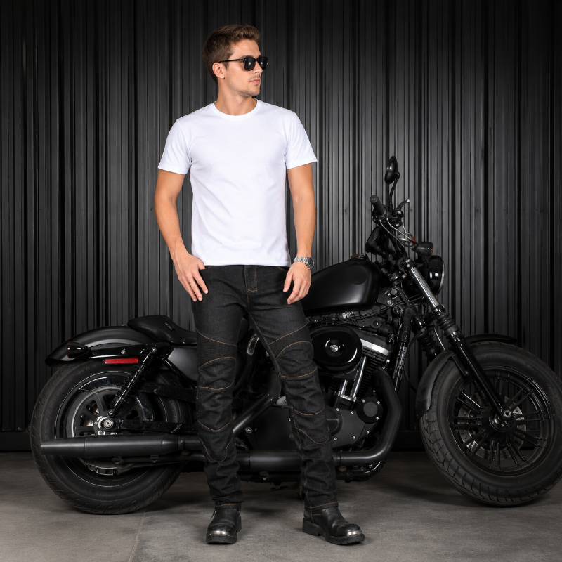 A man wearing a white crew-neck t-shirt and black denim jeans stands in front of a black cruiser motorcycle against a dark corrugated metal wall.