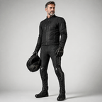 A man wearing a black leather motorcycle jacket, matching black pants with integrated knee and shin protection, black gloves, and riding boots, holding a black helmet against a light gray studio background.