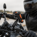 A close-up of a motorcycle handlebar mount holding a smartphone with an orange accent, installed on a motorcycle with a rider in a black helmet and leather jacket blurred in the background.