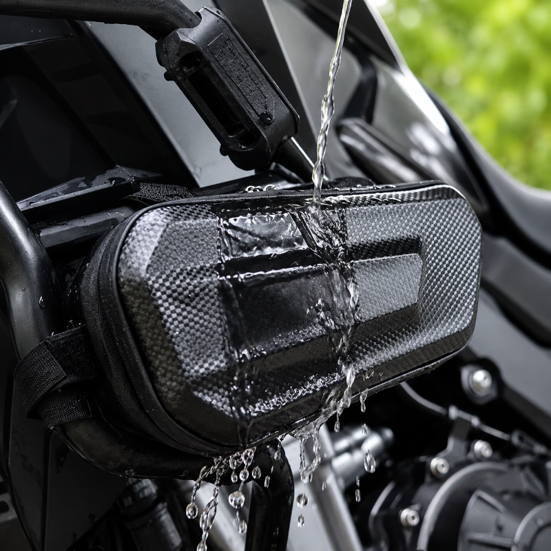 A black motorcycle tank bag with a carbon-fiber-look textured pattern, shown mounted on a bike and being splashed with water to demonstrate waterproofing.