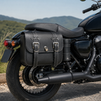 Black leather motorcycle saddlebags with silver buckles and a small lock, shown mounted on the rear of a black motorcycle outdoors with hills in the background.