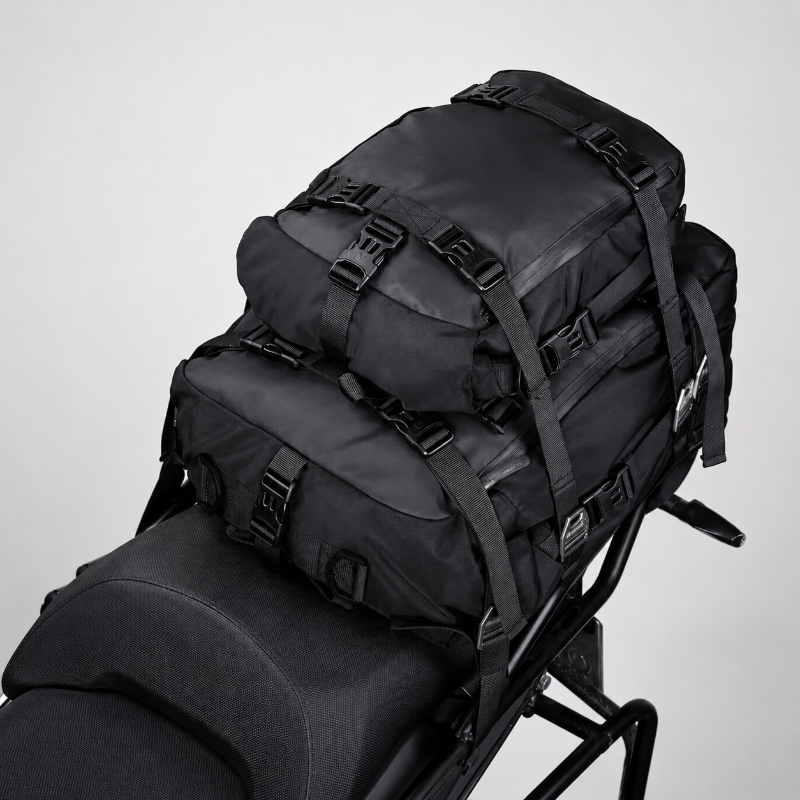 A black motorcycle luggage rack bag with multiple adjustable straps and buckles, shown mounted on a motorcycle seat against a light gray studio background.