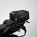 A black motorcycle luggage bag mounted on the rear rack of a black motorcycle, photographed against a plain light gray studio background.
