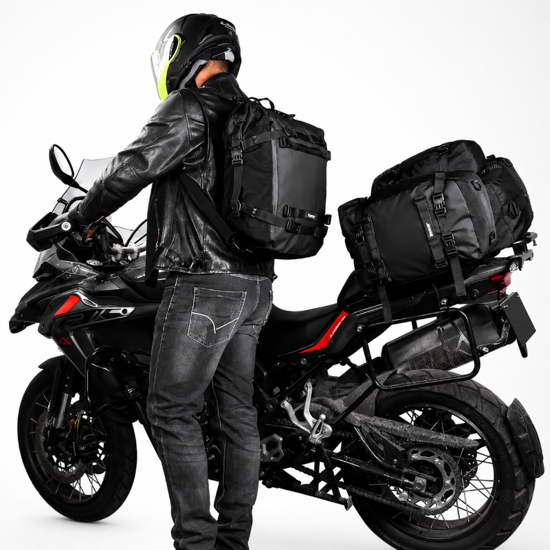 A black motorcycle touring backpack shown on a rider wearing a helmet and leather jacket, positioned next to a black motorcycle on a white studio background.