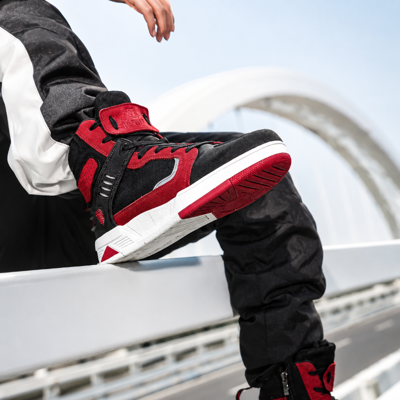 A close-up lifestyle shot of a person wearing a black and red high-top sneaker, sitting on a white railing with a blurred outdoor background.