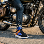 A person wearing a pair of black, blue, and orange high-top sneakers sits on a motorcycle on an asphalt road.