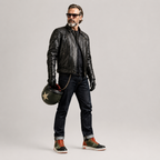 A man wearing a black leather jacket, dark jeans, black gloves, and sunglasses holds a black helmet with a white star on the left hand, standing against a light gray studio background.