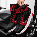 A pair of black and red high-top sneakers held by a hand in front of a blurred black motorcycle. The shoes feature a black and red color-blocked design with white midsoles.