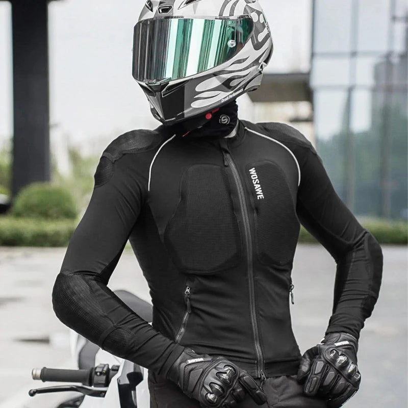 A person wearing a black full-face motorcycle helmet with a reflective visor, a black long-sleeve riding jersey with white piping and visible impact protection panels, black gloves, and a motorcycle in an outdoor urban setting.