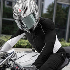 A person wearing a black and white riding jacket, black pants, and a white AGV full-face helmet with a clear visor, seated on a white Ducati motorcycle in an outdoor urban setting.