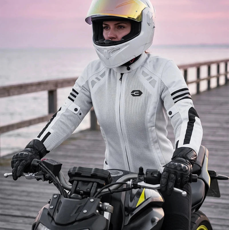 A woman wearing a white full-face motorcycle helmet and a white motorcycle jacket sits on a black motorcycle on a wooden boardwalk by the water at sunset.