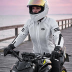 A woman wearing a white full-face motorcycle helmet and a white motorcycle jacket sits on a black motorcycle on a wooden boardwalk by the water at sunset.